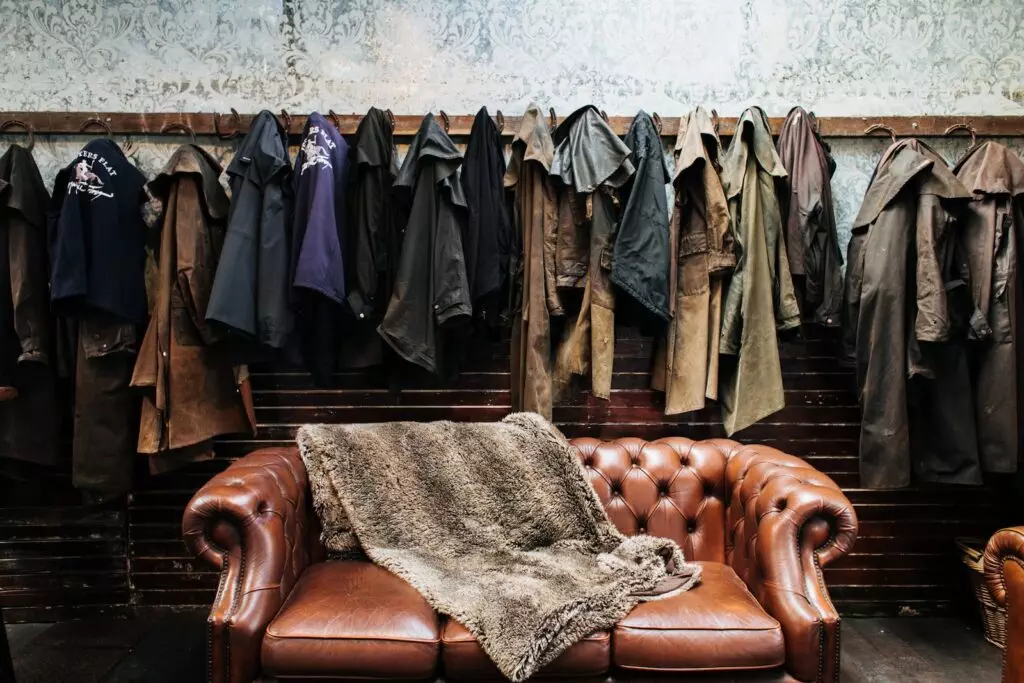 Various outerwear hanging on hooks at wall with wallpaper in light vintage studio with brown leather sofa with soft plaid